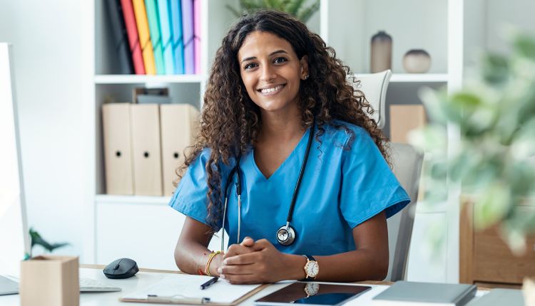 women wearing a stethascope in blue scrubs smiling at a desk with notes and tablet