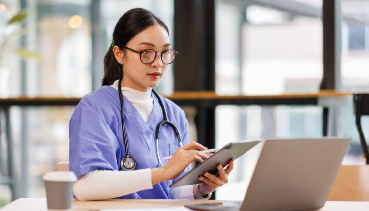 a medical assistant student in blue scrubs holds a tablet while sitting at a desk in front of a laptop computer